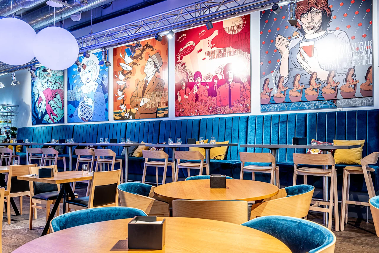 Stylish restaurant features blue velvet seating and wooden tables, surrounded by colorful pop art-style wall posters. Bright lighting accentuates the lively, modern atmosphere.