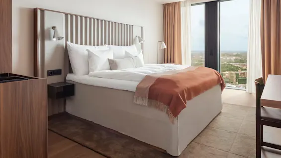 En snyggt bäddad säng med vita lakan och en orange filt står vid ett glasfönster med utsikt över stadslandskapet. Rummet har trämöbler och beige gardiner.