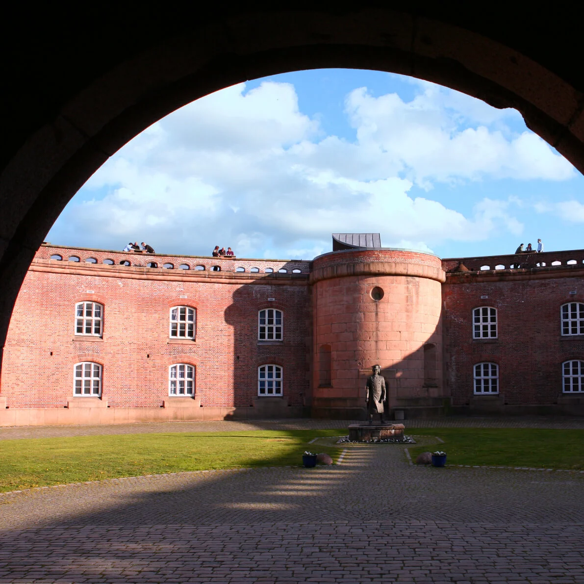 En buegang rammer inn en gårdsplass med en statue sentralt på brostein. Bygningen rundt har rød murstein og flere vinduer. Blå himmel og skyer er synlige over.