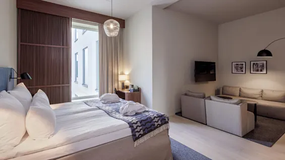 A modern bedroom with a neatly made bed, adjacent living area, and large window allowing natural light.