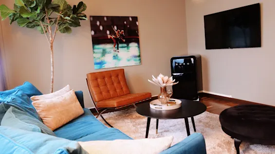 Blue sofa sits in a modern room with a brown chair, wall art, a television, a round table with a vase, and a small black fridge labeled "SMEG". A large plant is in the corner.