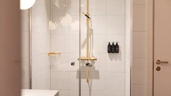 A modern shower with white tiles, gold fixtures, and toiletries, ready for a refreshing experience at Home Hotel Grand Bodø.