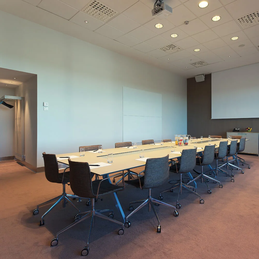 A long conference table is surrounded by chairs in a well-lit meeting room. A projector screen and a whiteboard are on the wall, with drinks on the table.