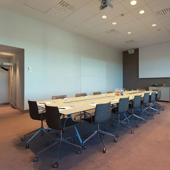 Ett långt konferensbord omges av stolar i ett väl upplyst mötesrum. En projektorduk och en whiteboard finns på väggen, med drycker på bordet.
