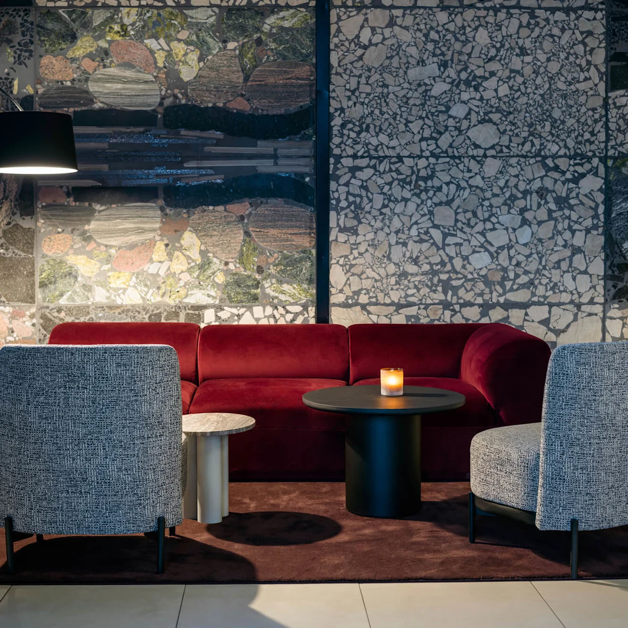 A red velvet sofa and two grey chairs surround a black round table with a candle. A patterned stone wall and floor lamp create a stylish, intimate lounge setting.