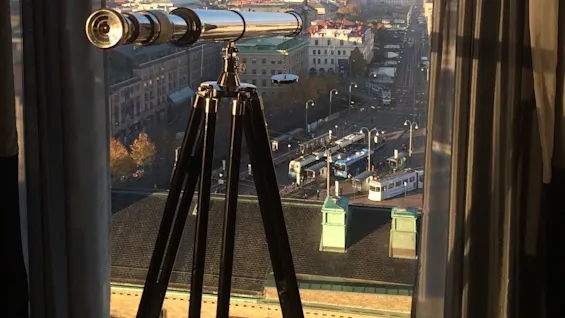 A telescope on a tripod stands by a window, overlooking a cityscape with buildings, roads, and trams under a clear blue sky.