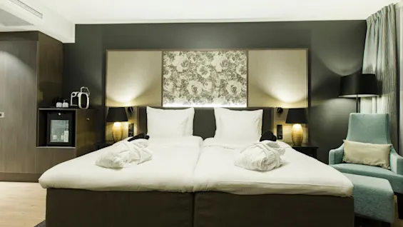 A neatly made double bed with white linens and two folded bathrobes, flanked by lamps, in a modern hotel room. A teal armchair and a minibar with a coffee machine are nearby.