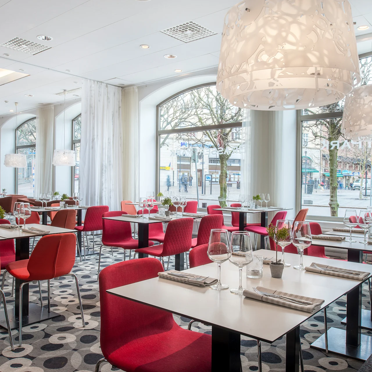 Bord med røde stoler er dekket med glass i en moderne restaurant, med store pendellamper og utsikt til trær og en bygate gjennom høye vinduer.