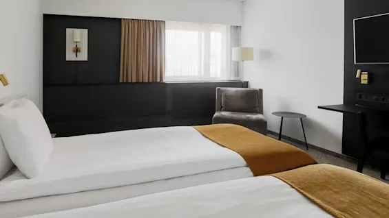 A hotel room features two neatly made beds with orange blankets. Nearby, a gray armchair sits beside a small round table and lamp. A TV is mounted on the black wall.
