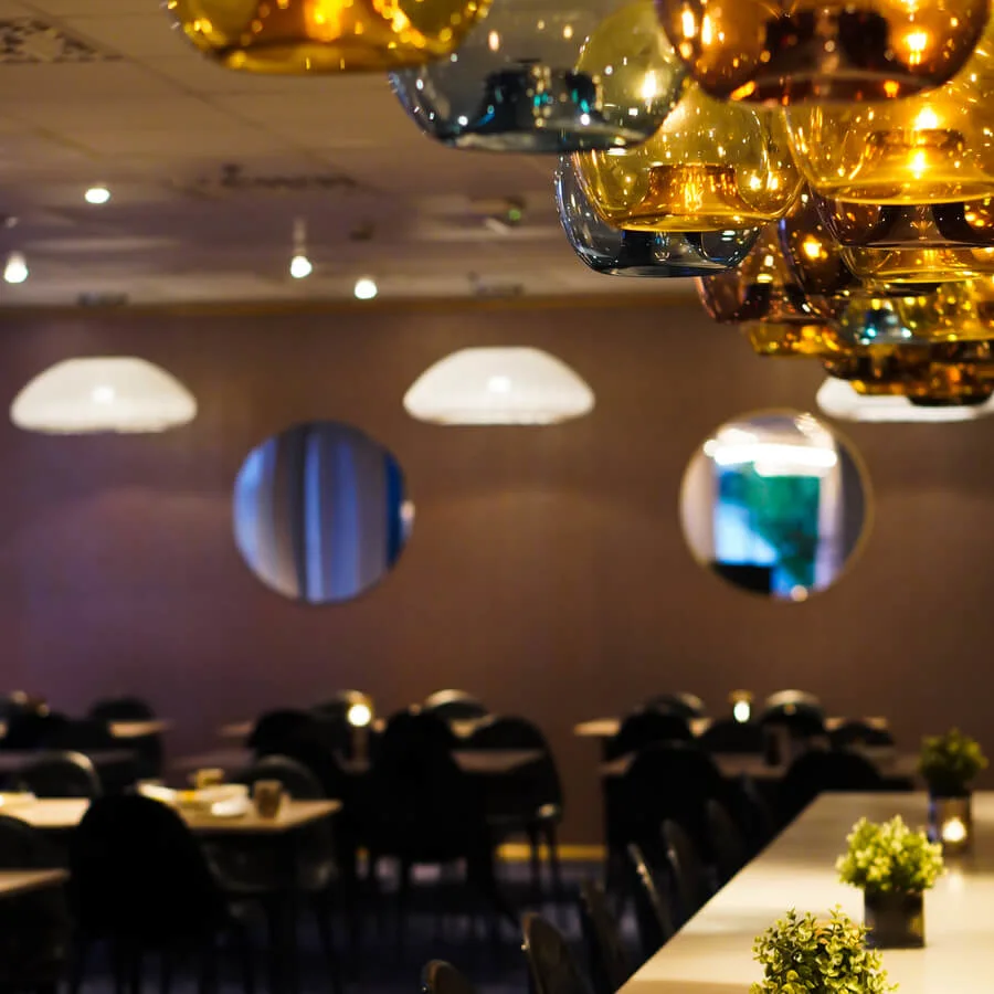 Pendant lights hang over a restaurant setting with tables and chairs. Soft lighting creates a warm atmosphere, with small plants and candles decorating the tables.
