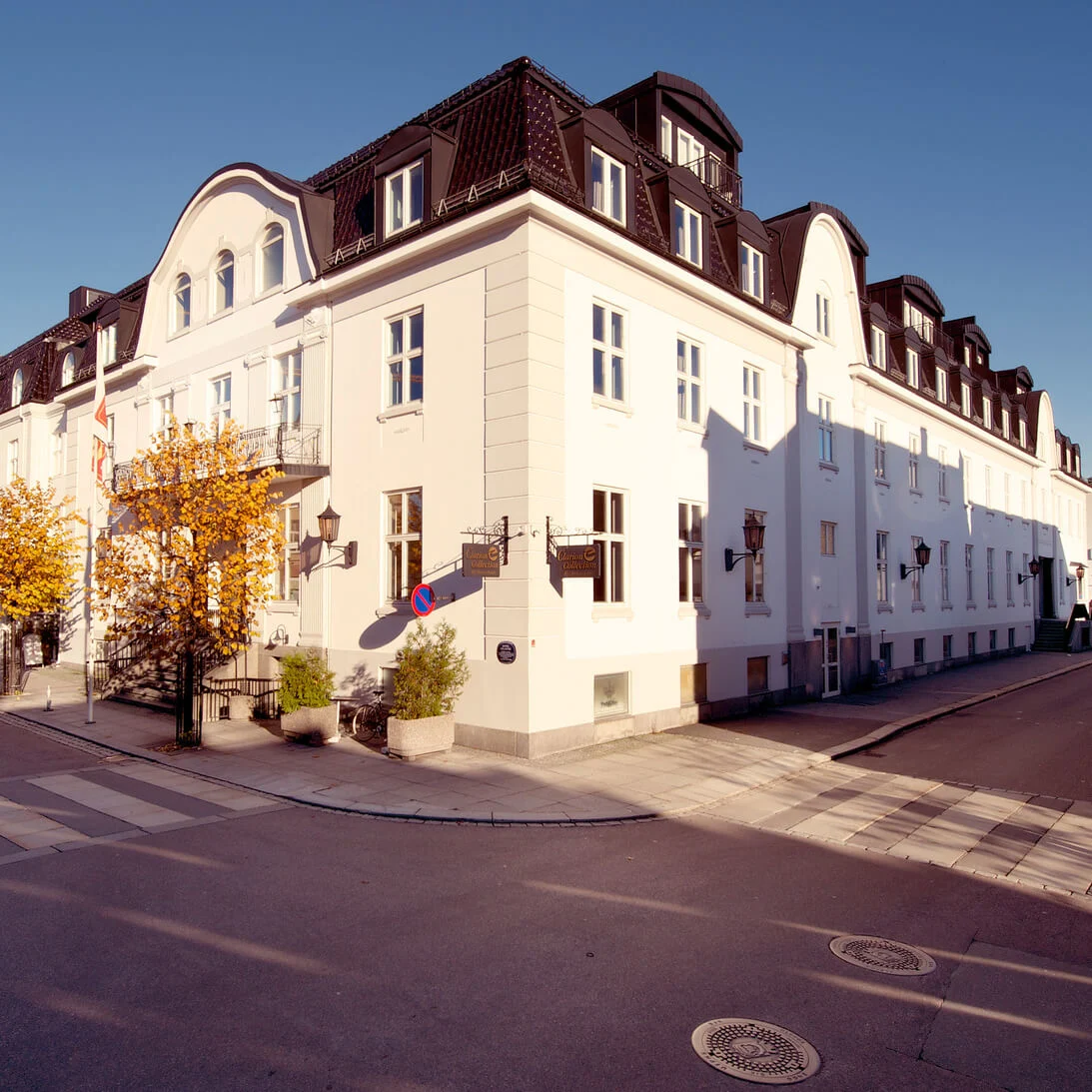 Ett vitt hörnhus med valvbågade fönster under en klarblå himmel vid en stadsintersection, med människor och fordon.