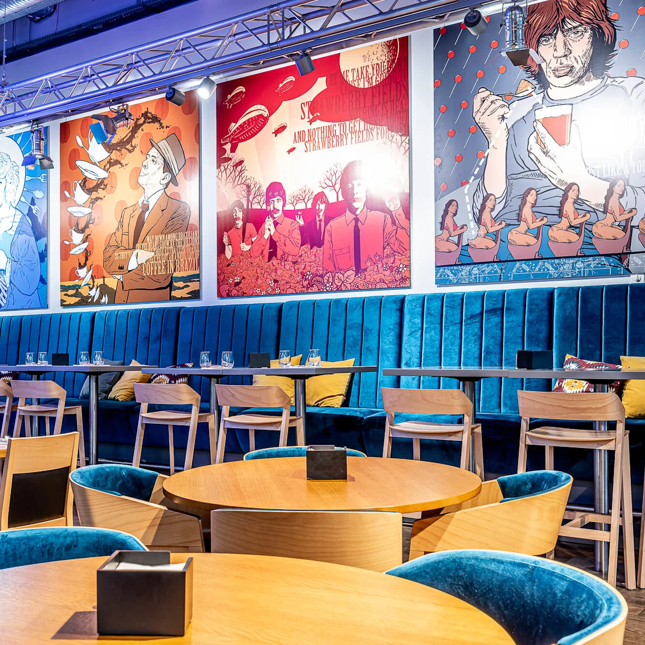 Trendig restaurang har blå sammetsstolar och träbord omgivna av färgglada väggaffischer i popkonststil. Ljusstark belysning betonar den livliga, moderna atmosfären.