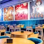 Stylish restaurant features blue velvet seating and wooden tables, surrounded by colorful pop art-style wall posters. Bright lighting accentuates the lively, modern atmosphere.