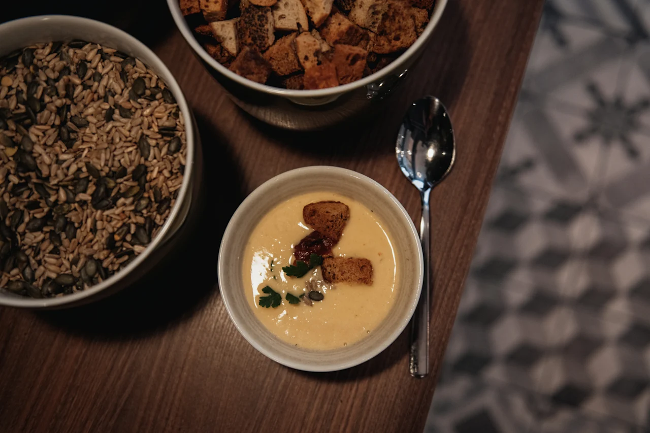 En bolle kremet suppe med krutonger og en skje, akkompagnert av boller med frø og flere krutonger på et trebord.