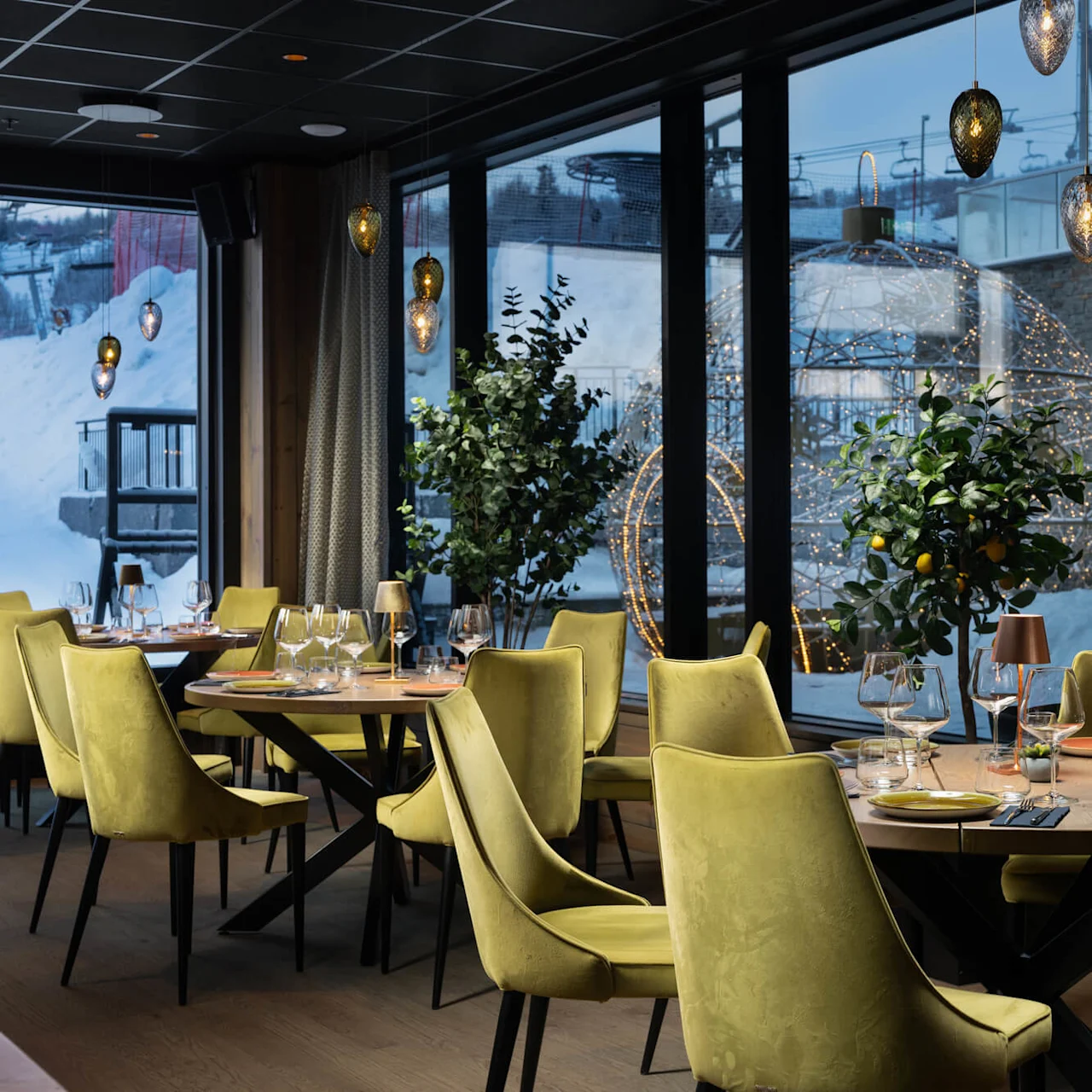 Bordene er dækket med glas og gule stole i en hyggelig, moderne restaurant. Store vinduer viser en sneklædt udendørs scene med dekorative lys.