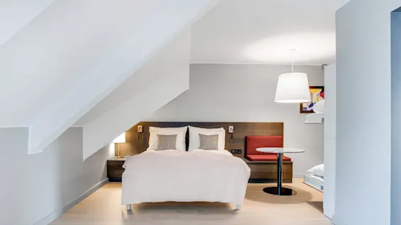 En seng med hvidt sengetøj og puder under et skråt loft, flankeret af to lamper og et lille bord med rød siddeplads. Moderne, minimalistisk soveværelsesdesign.