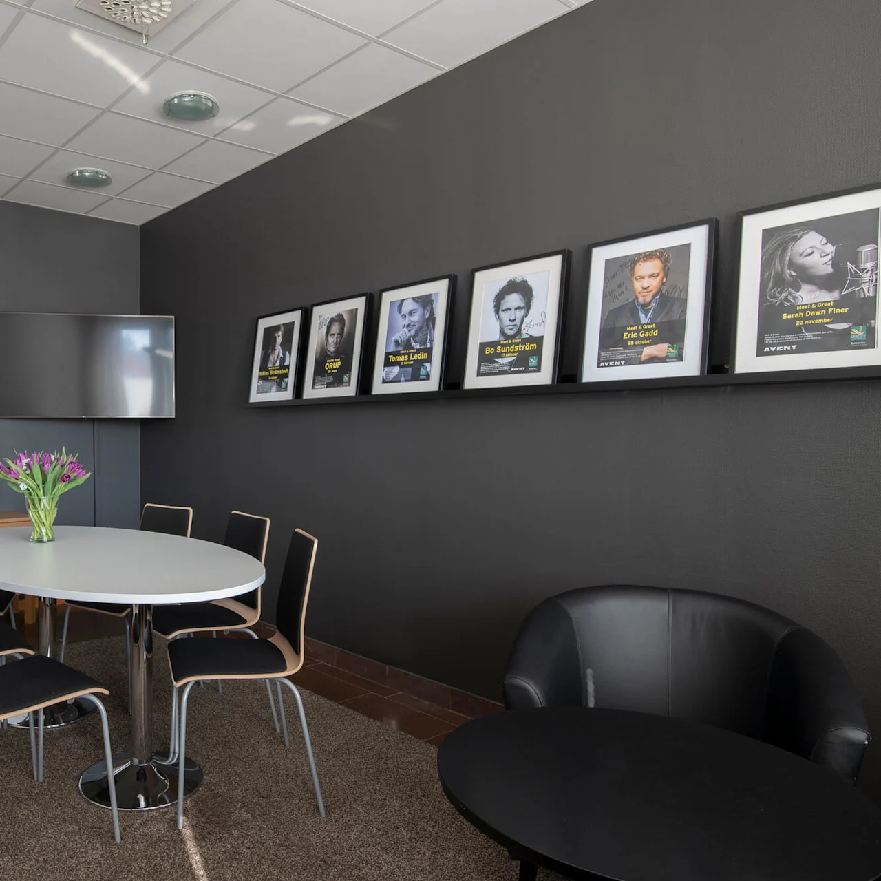 Runt bord omgiven av stolar, med en vas fylld med lila blommor, i ett konferensrum med whiteboard, TV och vägg med inramade kändisfoton. Text: "Orup, Tomas Ledin, Bo Sundström, Eric Gadd, Sarah Dawn Finer."
