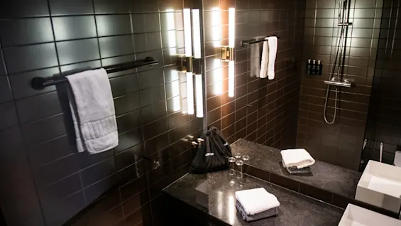 Håndklæder hænger på stænger i et moderne badeværelse med mørke fliser, med oplyste spejle, en bruser og en bordplade med foldede håndklæder og toiletartikler.