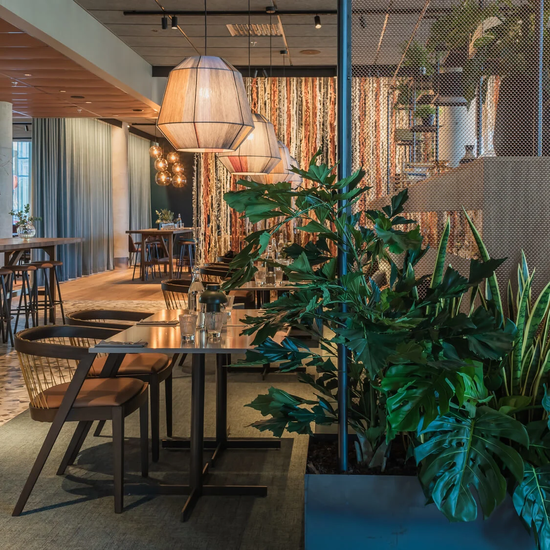 Lyxig restauranginteriör med moderna pendellampor som lyser upp träbord och stolar; frodiga gröna växter ger liv mot mjuk, varm belysning och skapar en inbjudande och stilren matmiljö.