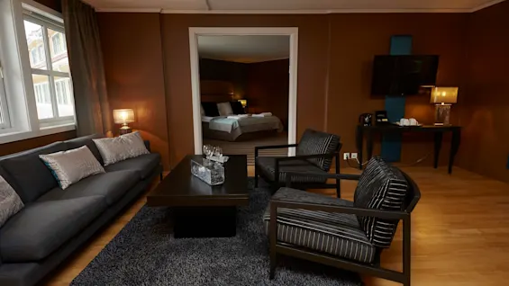 A gray couch and two black armchairs surround a dark coffee table on a shaggy rug, in a warmly lit living room adjoining a bedroom.