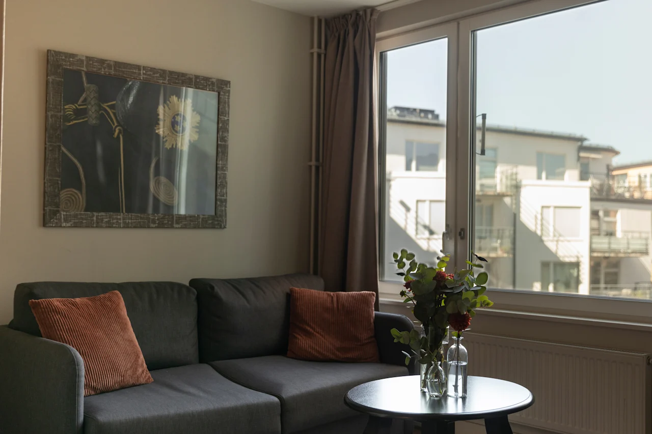 En bekväm hotellvardagsrumsdel med en soffa, ett soffbord med blommor och en ljus fönsterutsikt över byggnader.