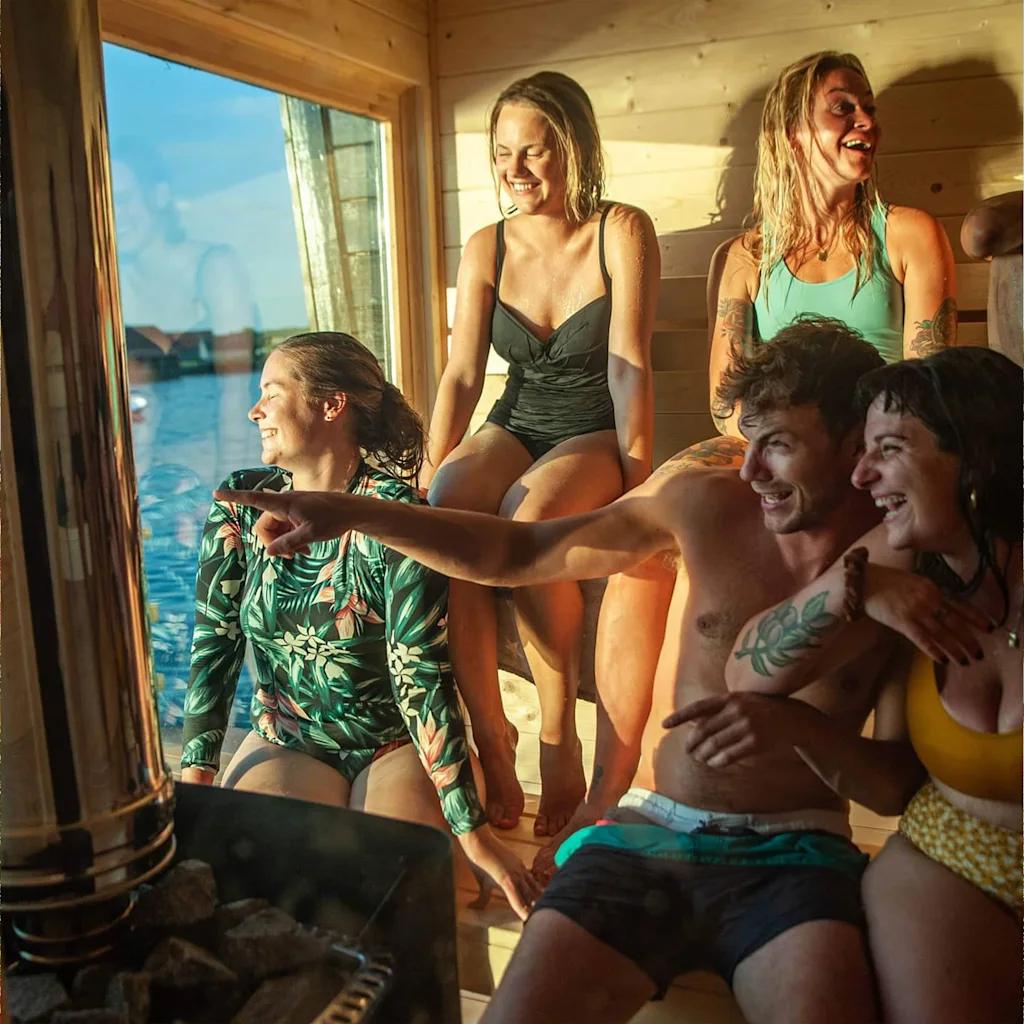 Ryhmä ystäviä nauraa ja osoittaa ikkunasta puusaunasta nauttien näkymästä veteen.