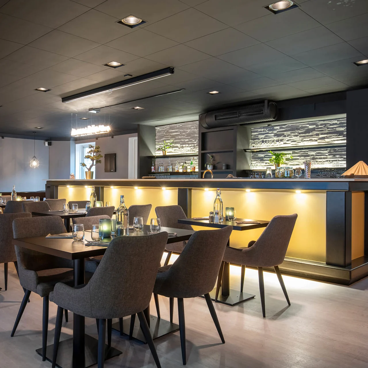 Ett modernt matrum har stoppade stolar runt mörka träbord med glas och ljus. I bakgrunden finns en elegant bar med upplysta hyllor.