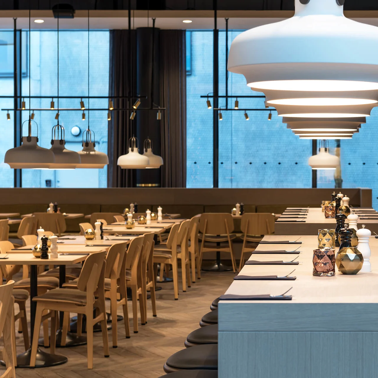 Bredt bord med servietter og krydder står under store hvite taklamper i en moderne restaurant, omgitt av stoler og med store vinduer i bakgrunnen.