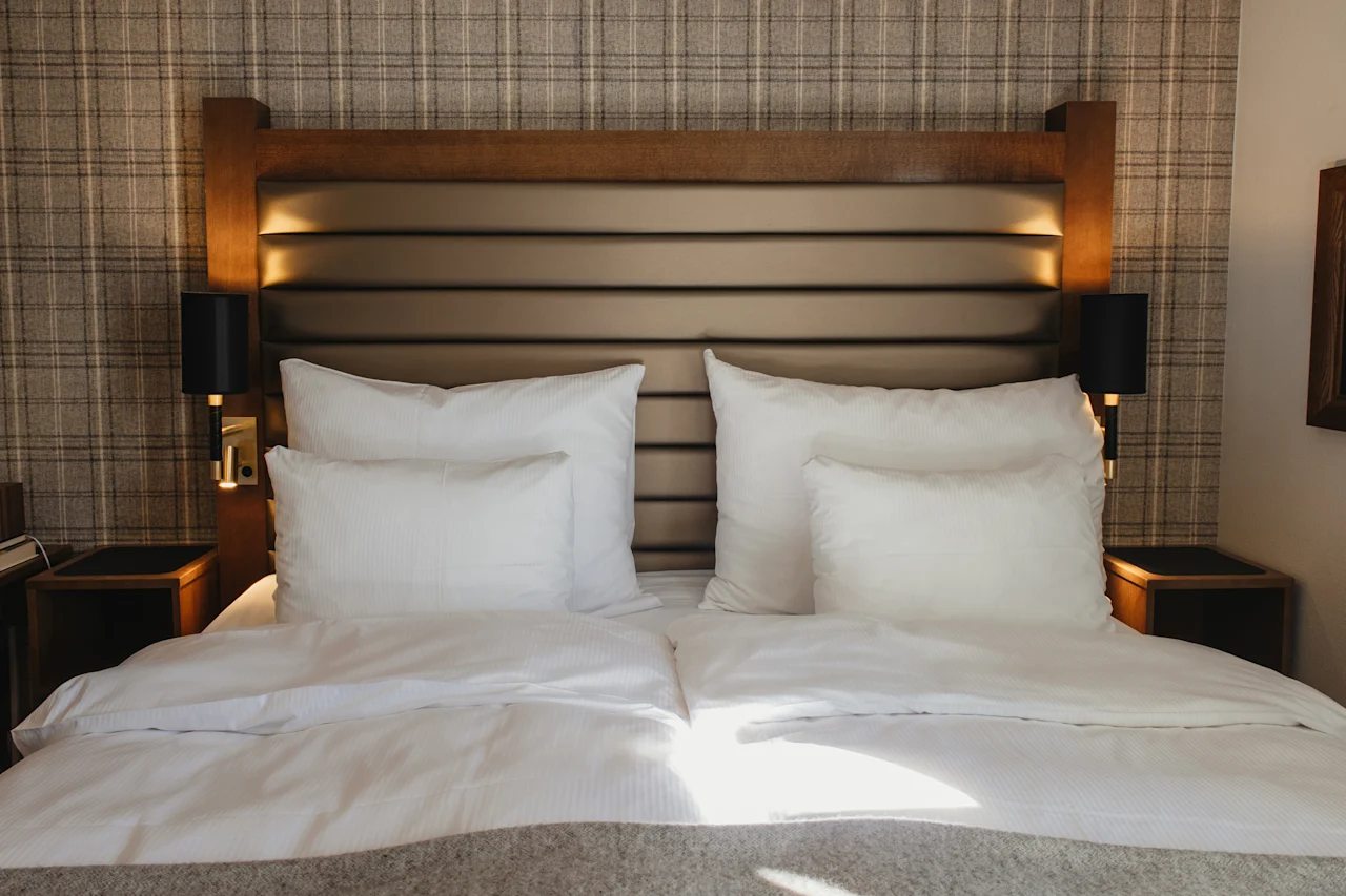 Et hyggeligt hotelværelse med en pænt redt seng, hvidt sengetøj og en stilfuld sengegavl. Sollys strømmer ind over sengen.