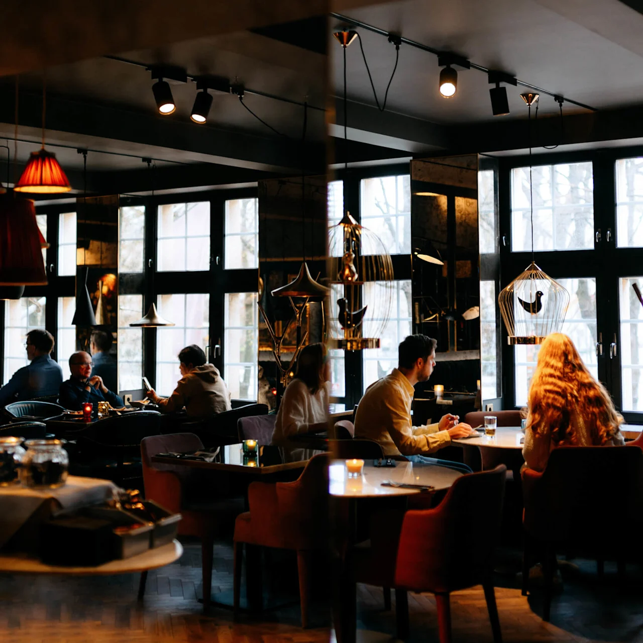 People sit at tables talking and dining in a warmly lit café with large windows, hanging lamps, and decorative cages. The ambiance is cozy and inviting.