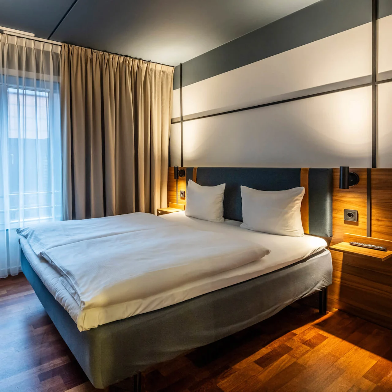 A modern hotel room featuring a double bed covered with white linens. The walls have wood paneling details, and the bed is flanked by reading lights and small bedside tables. Large curtains frame the window, allowing natural light to enter.