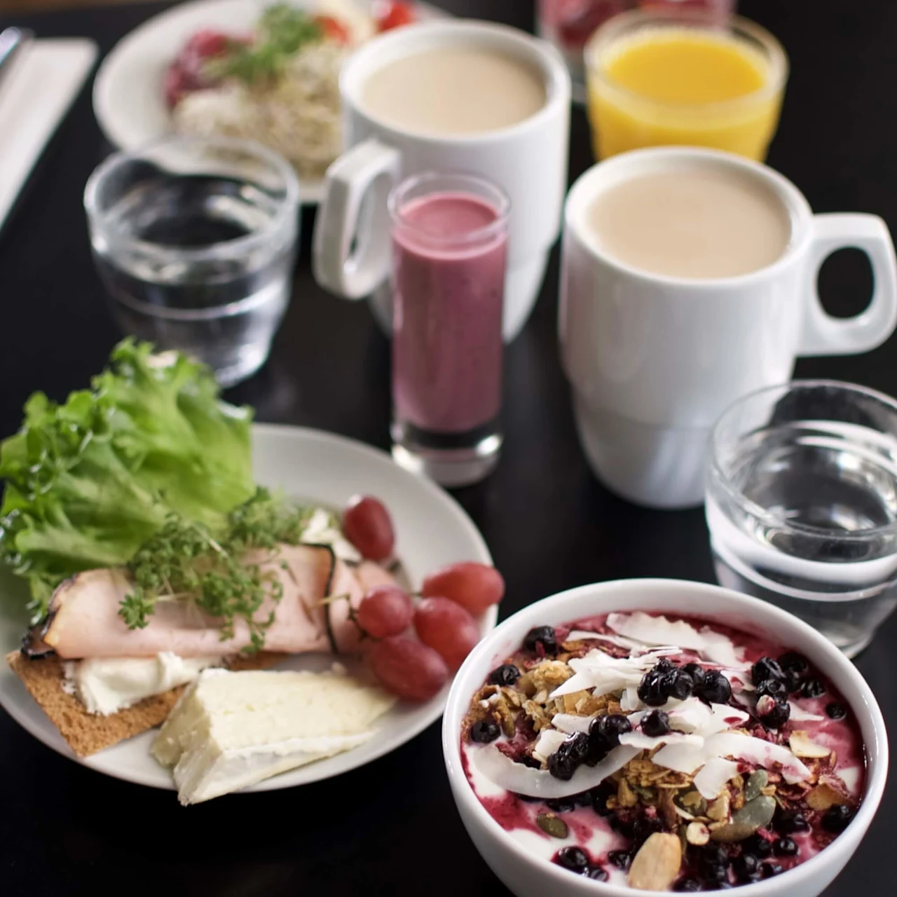 Yoghurt toppet med granola og bær står på et bord. Nær er tallerkener med brød, ost, druer, salat og drikker i krus og glass.