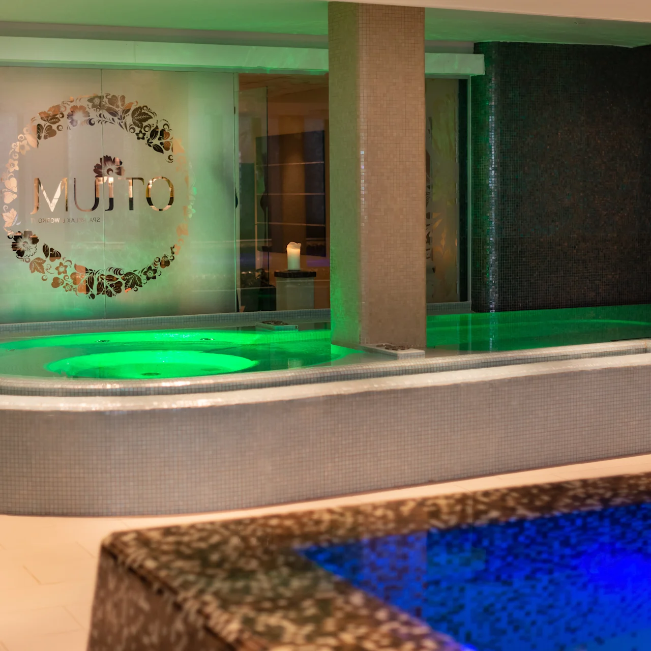 A circular pool emits green light, surrounded by a modern, tiled area. Wall displays the word "MUTTO" within a decorative circle. Calm, ambient lighting and design create a spa-like atmosphere.