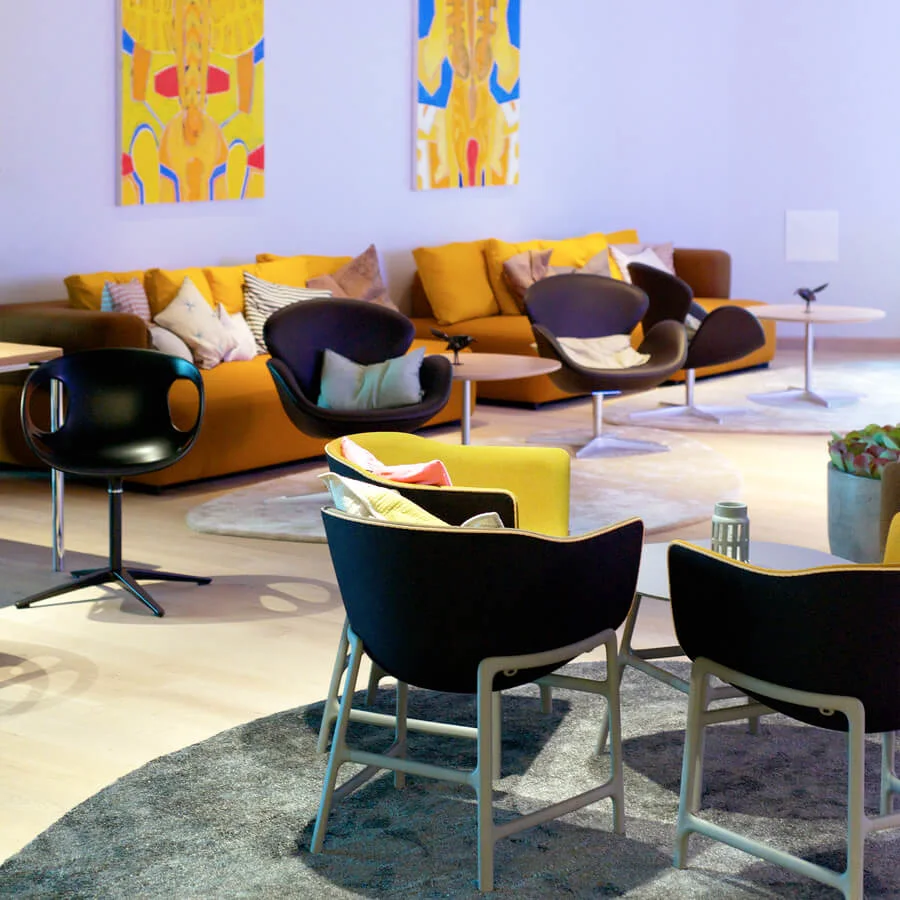 Modern lounge som har svarta och gula stolar med kuddar, placerade runt små bord på ett ljust golv. Ljusa abstrakta målningar hänger på en blek vägg och förstärker den mysiga atmosfären.