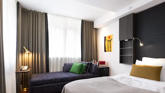 Et hyggeligt hotelværelse med en mørkeblå sofa og grønne puder ved siden af en seng. En gulvlampe oplyser området, med kunst og et lille skab i nærheden.
