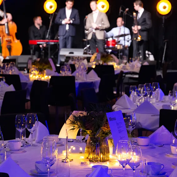 Et band spiller på scenen mens gjester spiser ved elegant dekkede bord med myk belysning på et kveldsarrangement. Meny synlig.