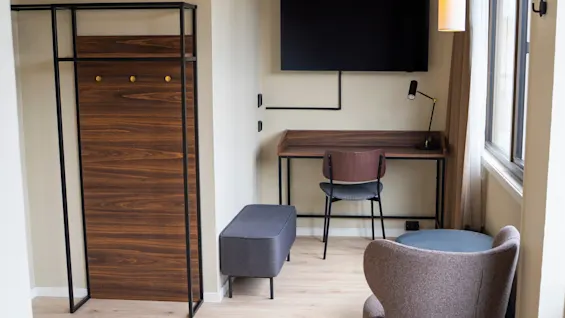 A modern hotel room with a desk, chair, television, a cozy armchair, and a coat rack, offering a comfortable stay.