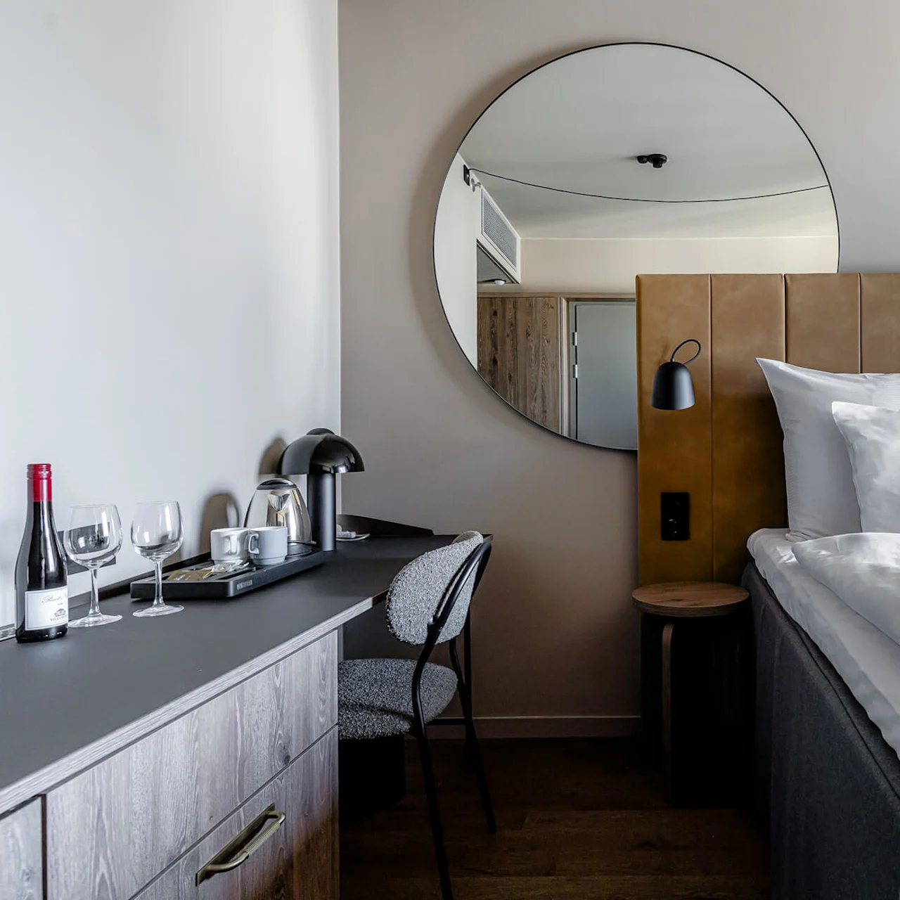 A neatly made bed with white pillows and a gray blanket sits beside a desk with a chair, a lamp, wine, glasses, and a coffee set, in a modern room.