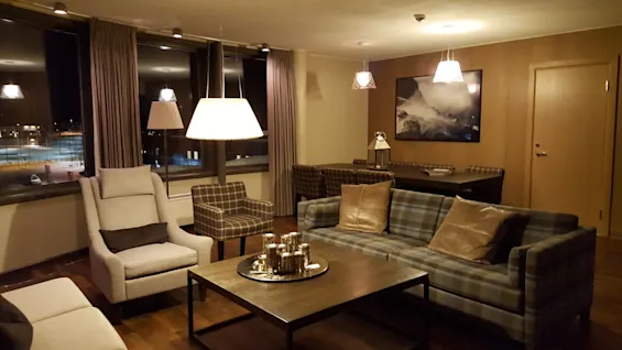 Et koselig oppholdsrom har en rutete sofa med puter, et sentralt kaffebord og en lenestol; myk belysning fra lamper lyser opp kunst på trevegger og et stort vinduutsikt.