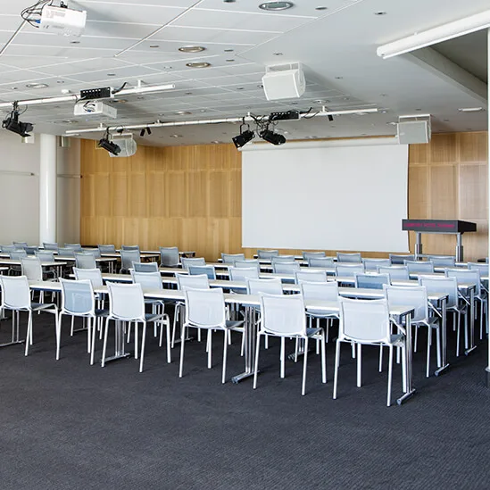 An empty conference room with rows of white chairs and tables facing a large projector screen. The room has a carpeted floor, white walls, and ceiling-mounted equipment.