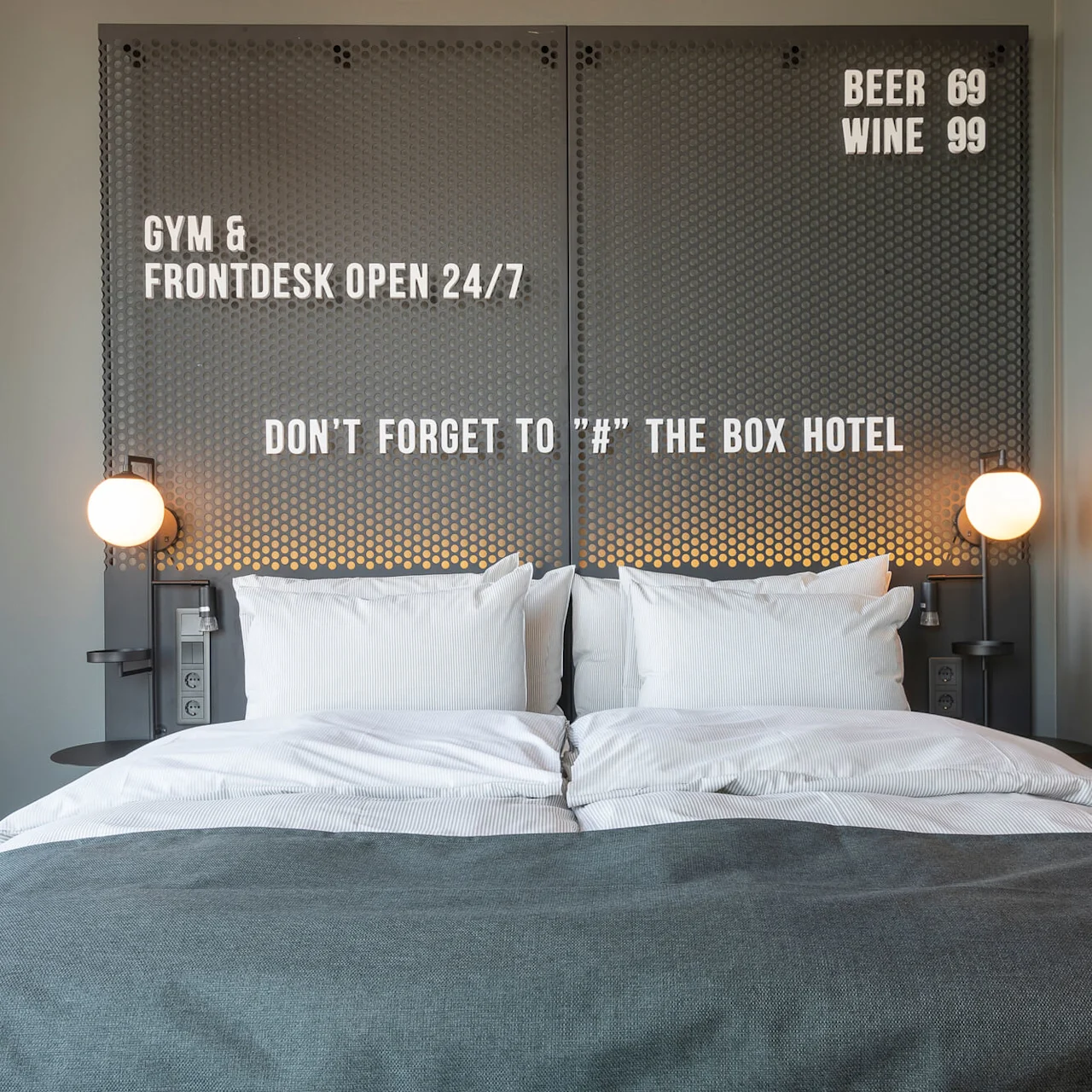 A neatly made double bed with white pillows and a dark blanket is flanked by wall lamps. Text: "GYM & FRONTDESK OPEN 24/7," "DON'T FORGET TO '#' THE BOX HOTEL," "BEER 69," "WINE 99."