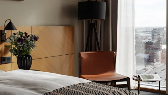 Et koselig hotellrom med en seng, blomster, en komfortabel stol og et stort vindu med utsikt over byen.