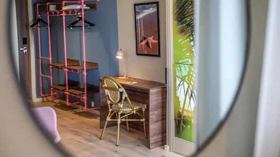 A wooden desk with a wicker chair faces a wall in a modern room. A table lamp and plant poster provide context, with pink pipe shelving visible nearby.