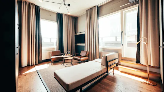 En moderne stue med en chaiselong, to brune stoler og et rundt bord med glass. Nøytrale gardiner rammer inn store vinduer, og en moderne taklampe tilfører eleganse.
