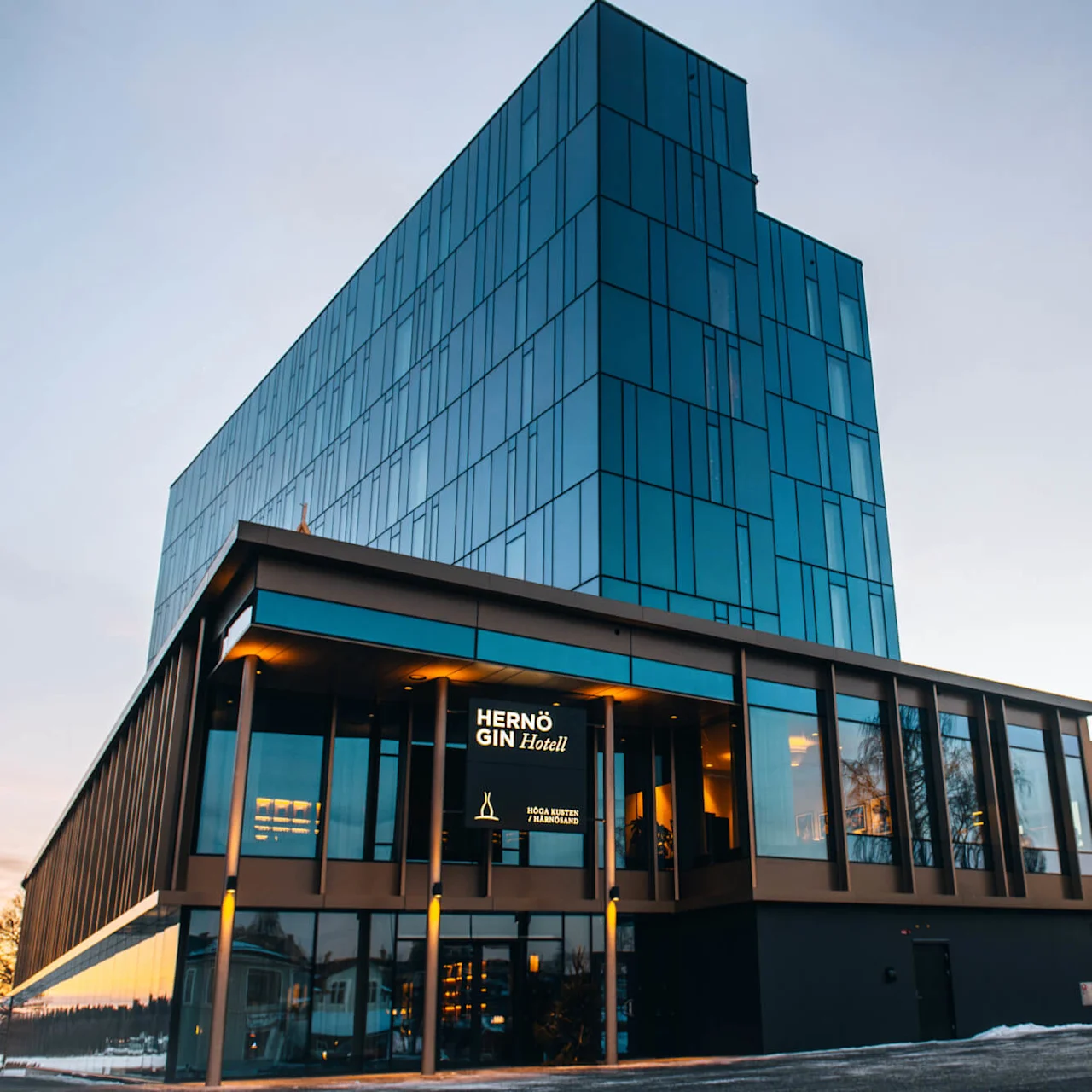 En modern glasbyggnad med blå fönster står i skymningen; en upplyst skylt lyder "HERNÖ GIN Hotell," omgiven av snötäckt mark och avlägsna träd under en blek himmel.