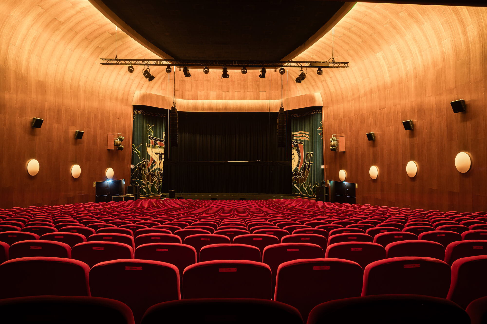 Rows of red theater seats face a stage with closed curtains in a warmly lit, wooden-walled auditorium, creating an inviting atmosphere for a performance.