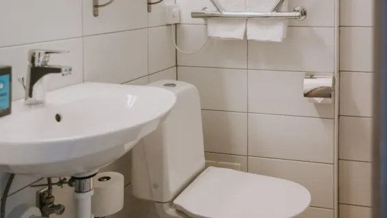 Et hvidt toilet og håndvask i et flisebelagt badeværelse. To glas og en sæbedispenser på en hylde, håndklæder hænger på en håndklædetørrer.