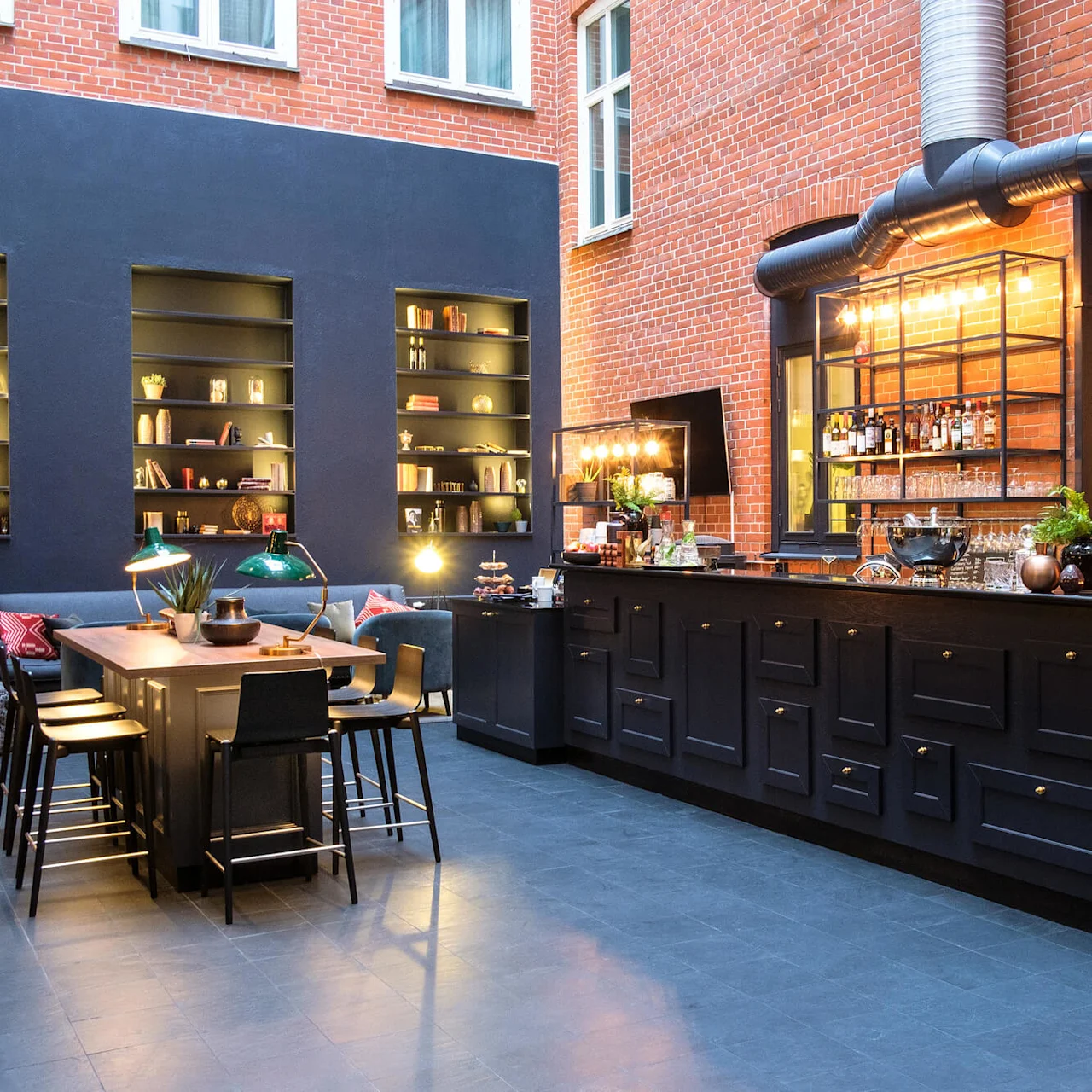 An elegant indoor bar area with stools, shelves with books and bottles, set against exposed brick walls and dark decor.