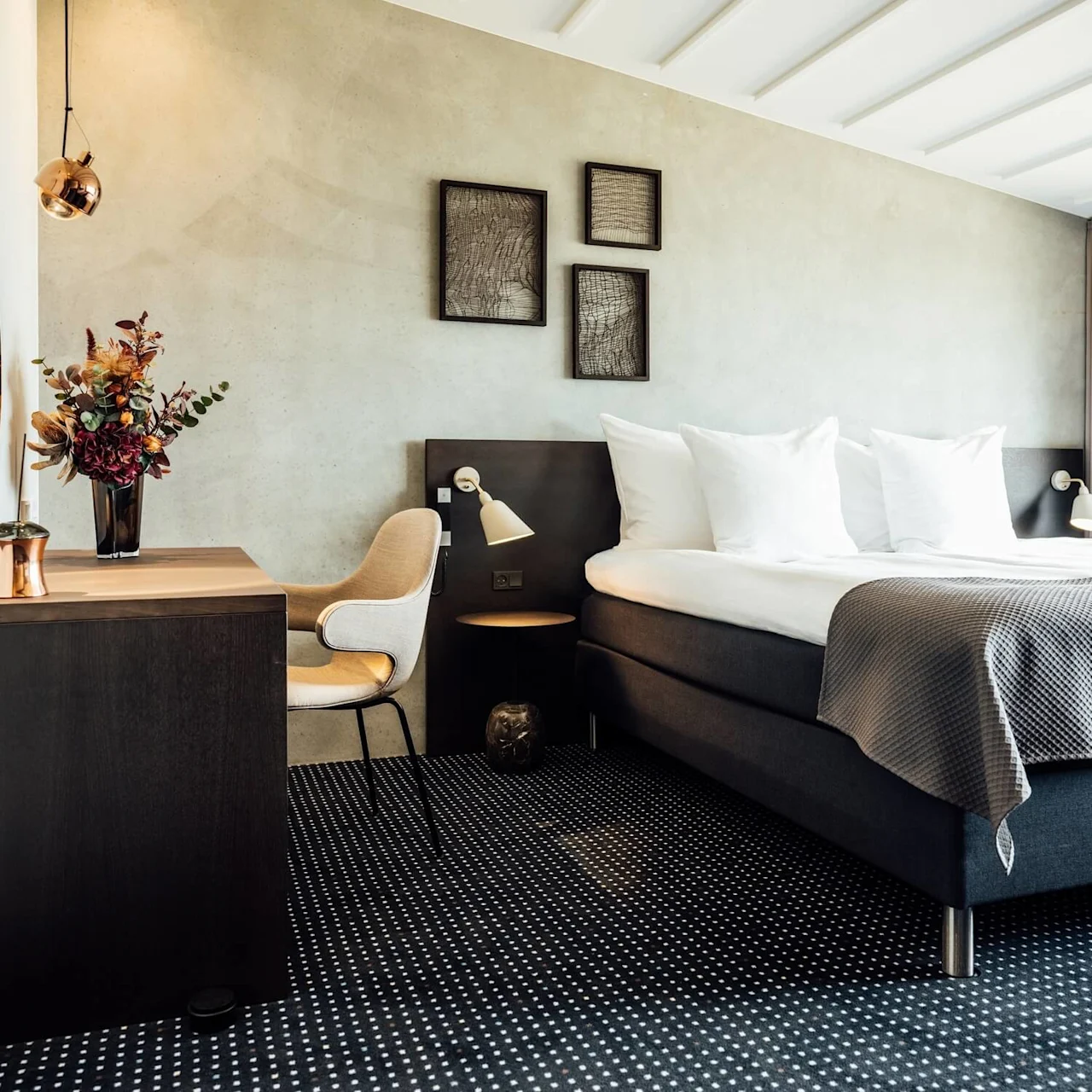 Et moderne hotelværelse har en pænt redt seng med hvide puder, ledsaget af et skrivebord med en vase med farverige blomster. Grå toner dominerer den rolige og elegante indstilling.