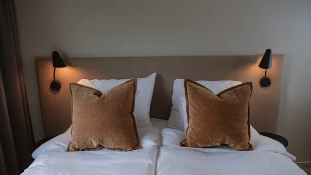 En prydligt bäddad säng har två stora bruna kuddar på vita sängkläder. Mjuk belysning från vägglampor skapar en mysig atmosfär i ett minimalistiskt sovrum med beige väggar.